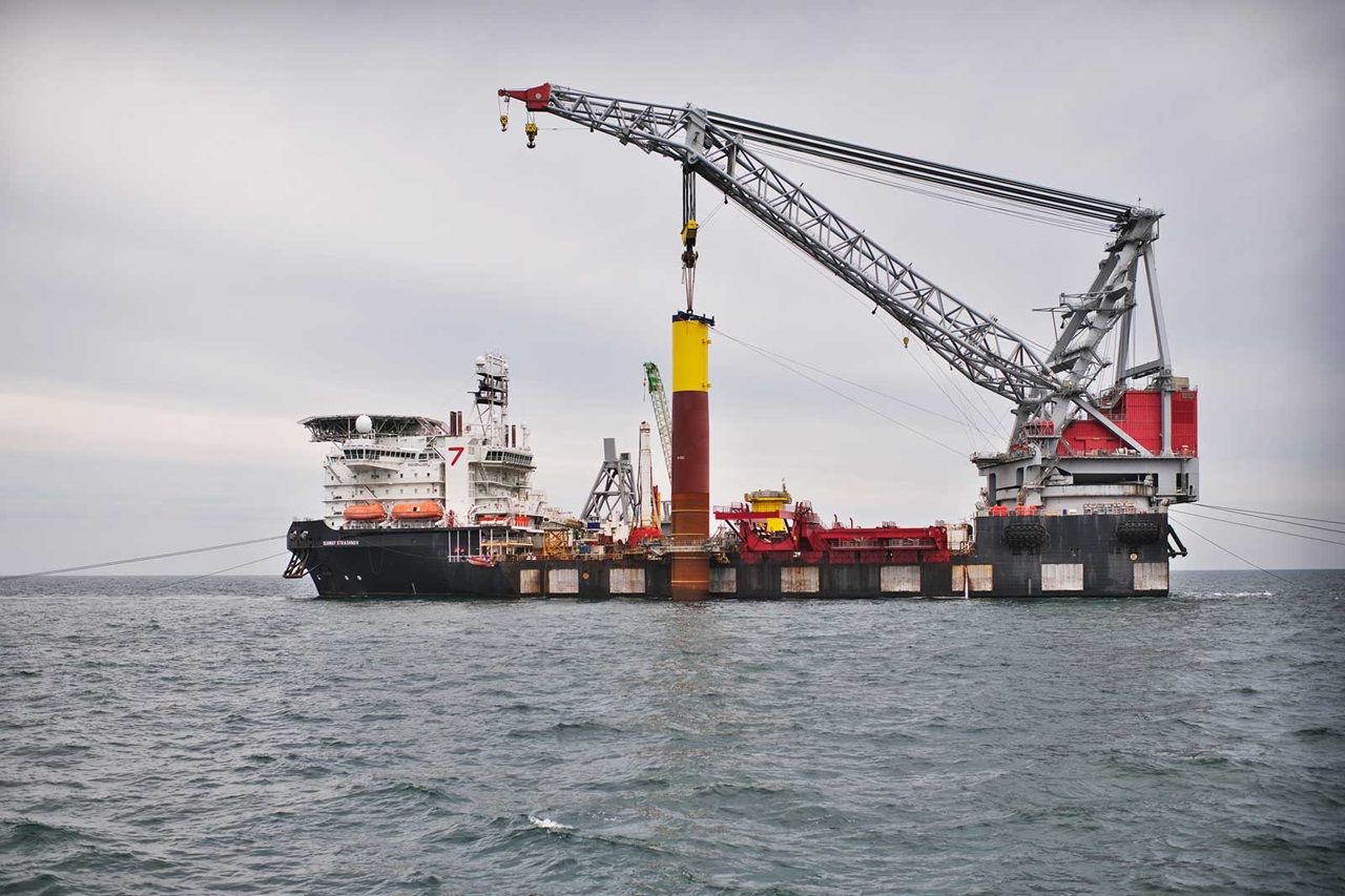 An offshore vessel with a large crane is lifting a yellow and brown pile over the water.