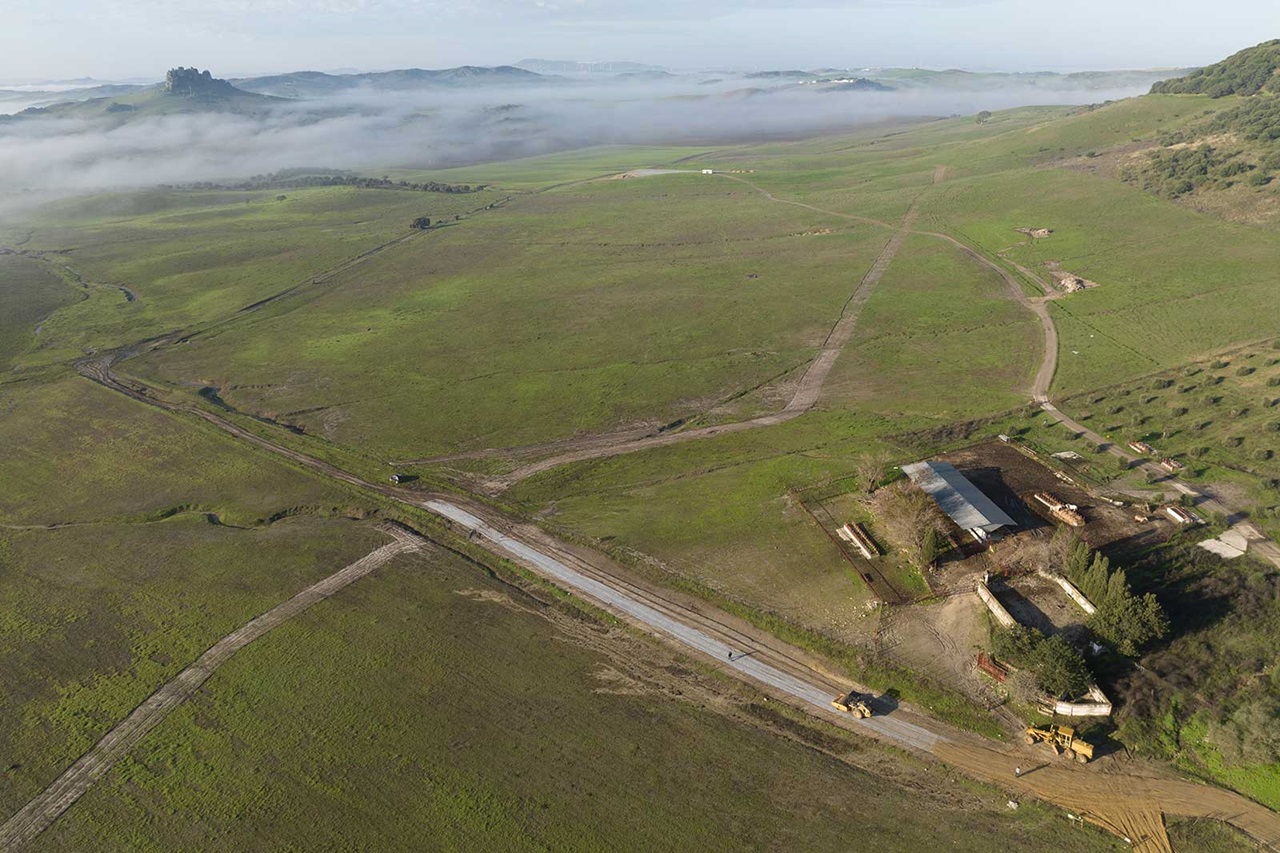 Aerial view of a green landscape with mist, featuring a barn, roads, and distant hills.