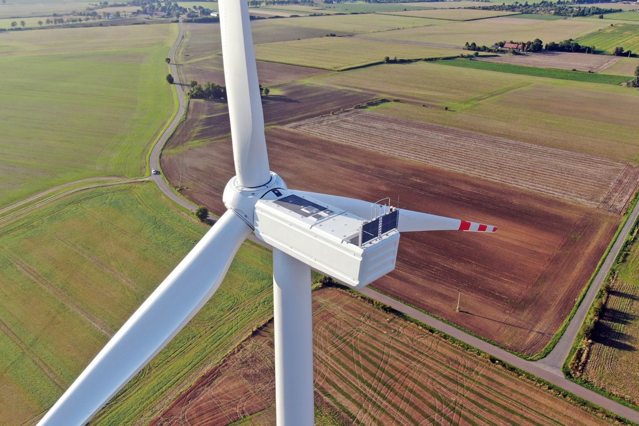 Aerial view of a large wind turbine with a white tower and three blades, set against a patchwork of green and brown fields.