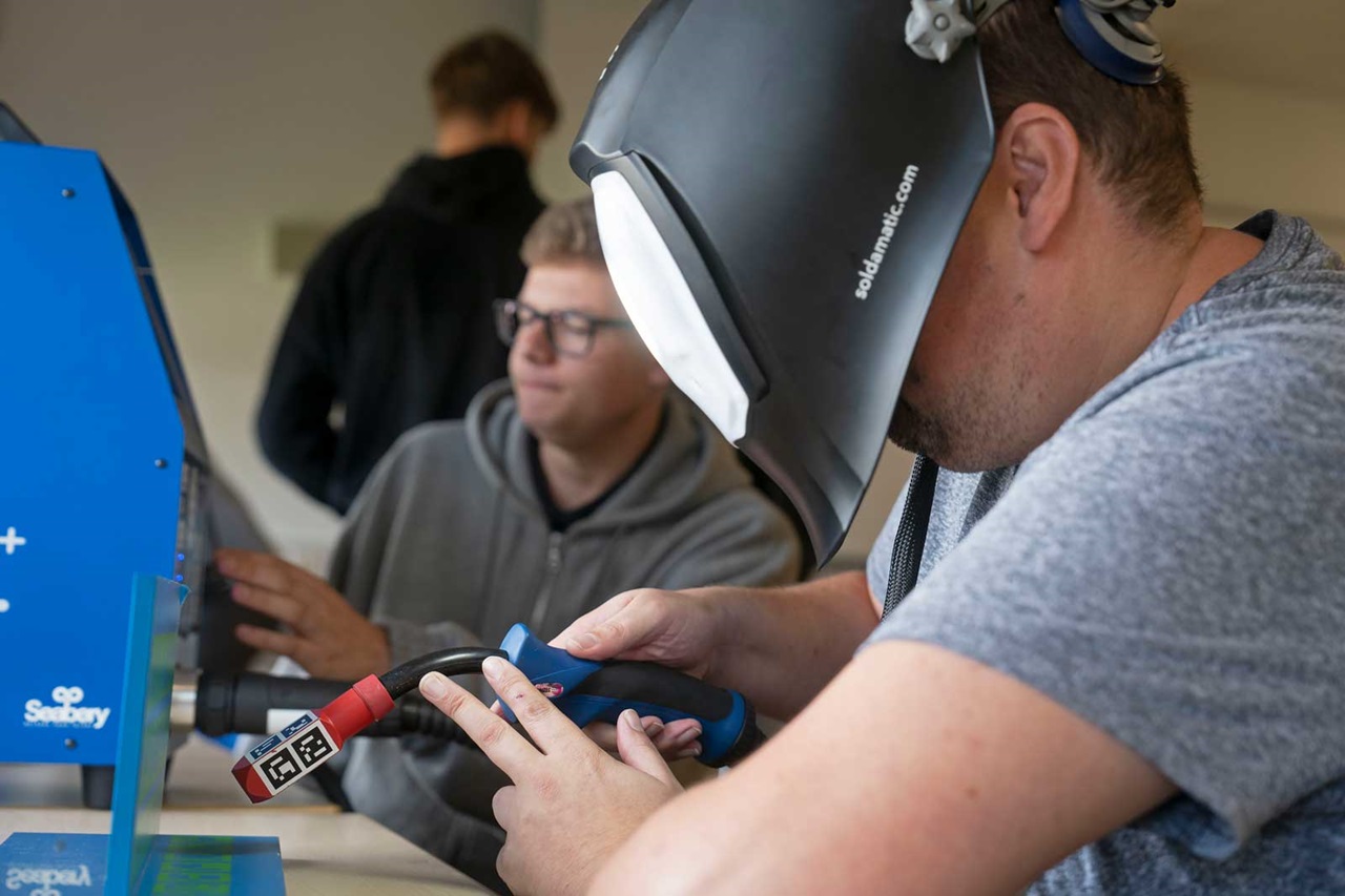 A man is wearing a welding helmet and holding a welding torch. Other people are visible in the background.