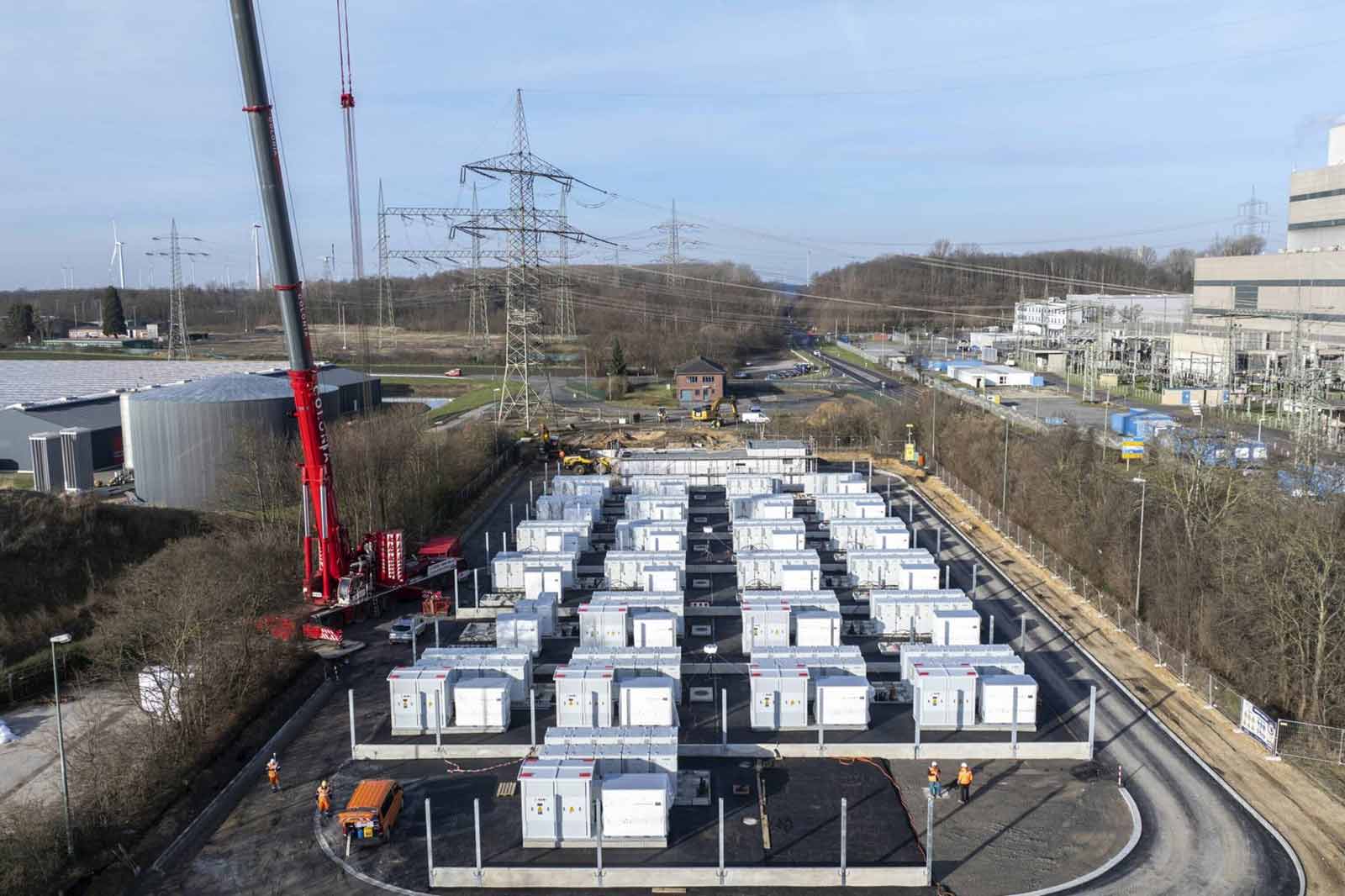Eine Baustelle mit vielen weißen Containern im Vordergrund, im Hintergrund Hochspannungsmasten und ein Industriegebäude.