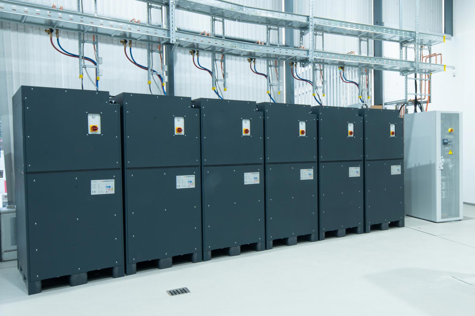 Sechs große, graue Energiekästen stehen nebeneinander, mit einem kleinen Steuergerät rechts daneben.