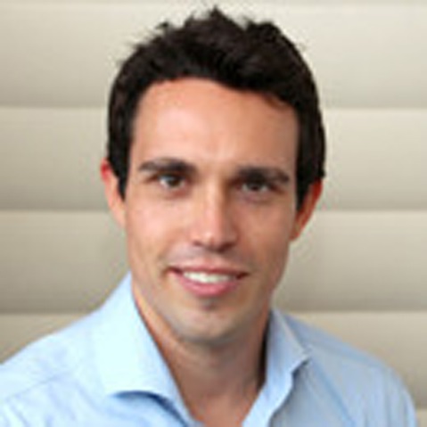 A man is wearing a blue shirt and standing in front of a light, textured wall.