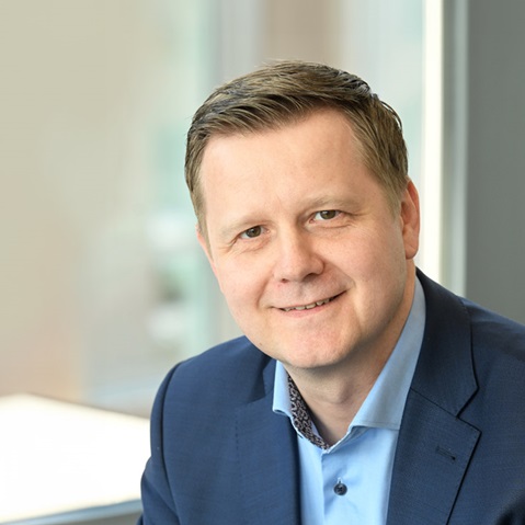 Ein Mann in einem blauen Anzug sitzt in einem modernen Büro mit großen Fenstern. Er trägt ein hellblaues Hemd und ein elegantes Muster. Portrait von Martin Modzelewski.