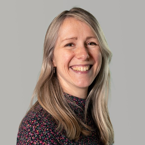A person with long blonde hair wears a patterned top and stands in front of a modern glass wall. Portrait of Geertje van Duijne.