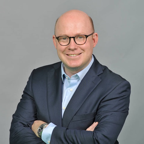 A man in a dark suit stands with crossed arms against a grey background. Portrait of Felix Christoph Ringenberg.