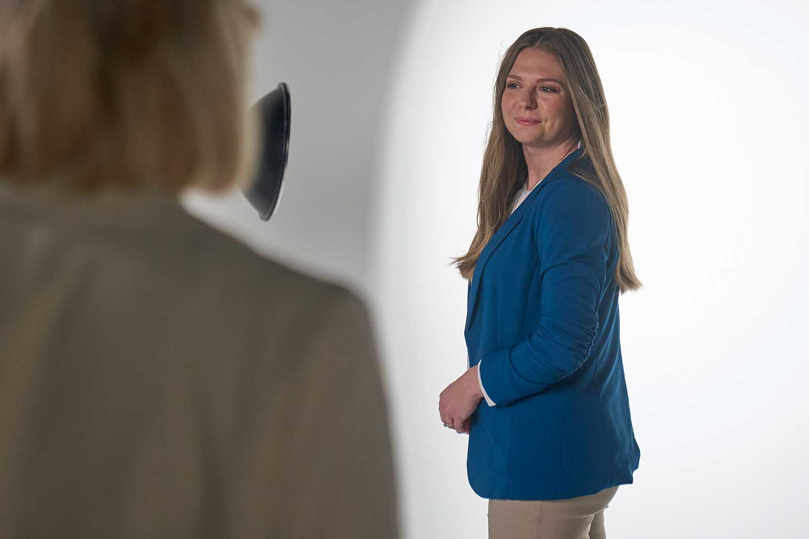 Eine Person mit langen, braunen Haaren trägt eine blaue Jacke und steht vor einem hellen Hintergrund.