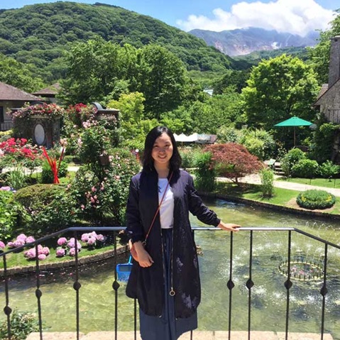 A person stands by a railing in a blooming garden with mountain views and a small pond in the background.