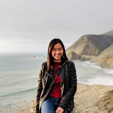 A person sits by the shore, wearing a black leather jacket and looking at the sea, with mountains visible in the background.