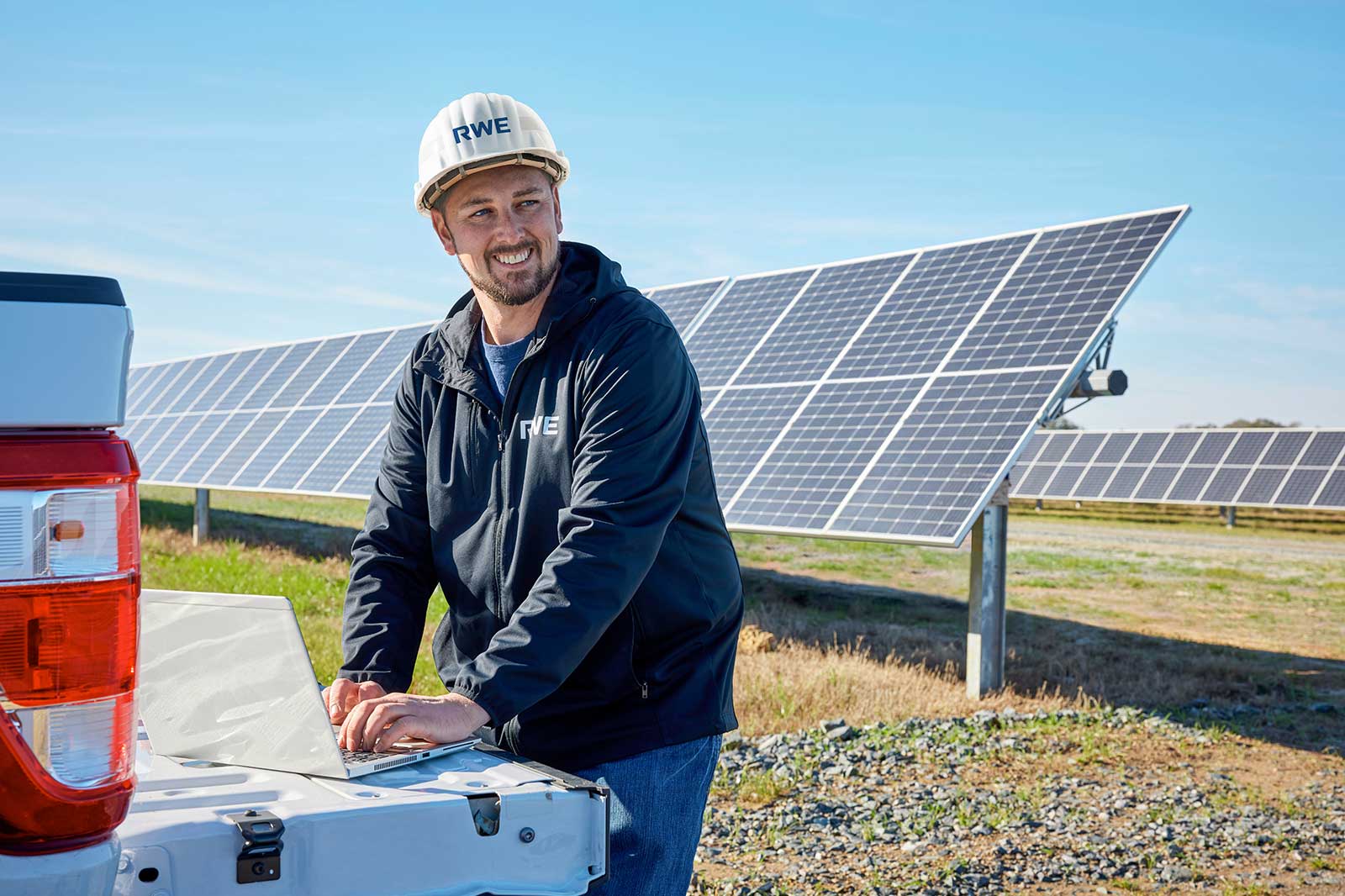 An RWE employee stands at his white pick-up truck and uses a laptop. In the background are solar panels on a meadow.