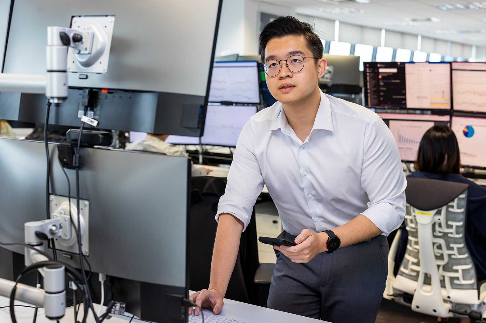 A man in a white shirt stands at a desk with multiple computer monitors displaying data charts and graphs.