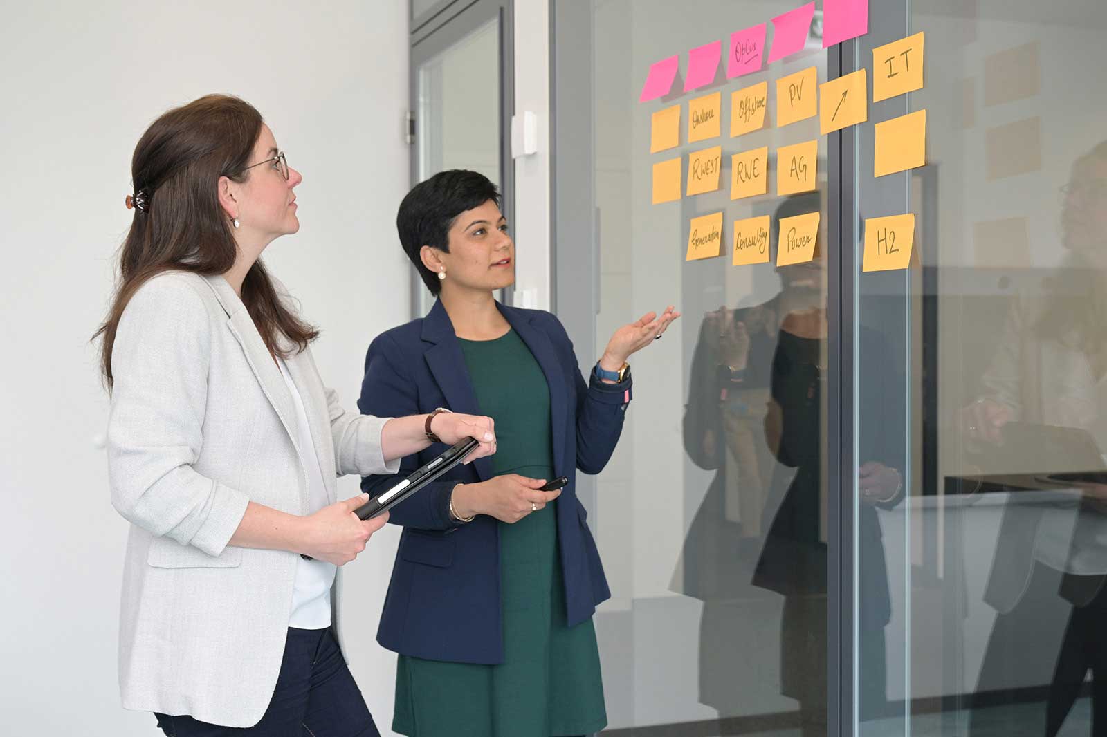 Two women discuss at a glass wall covered with colourful sticky notes, highlighting topics for a meeting.
