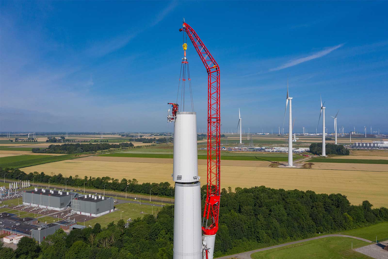 A crane lifts wind turbine components, surrounded by fields and additional wind turbines in the background.
