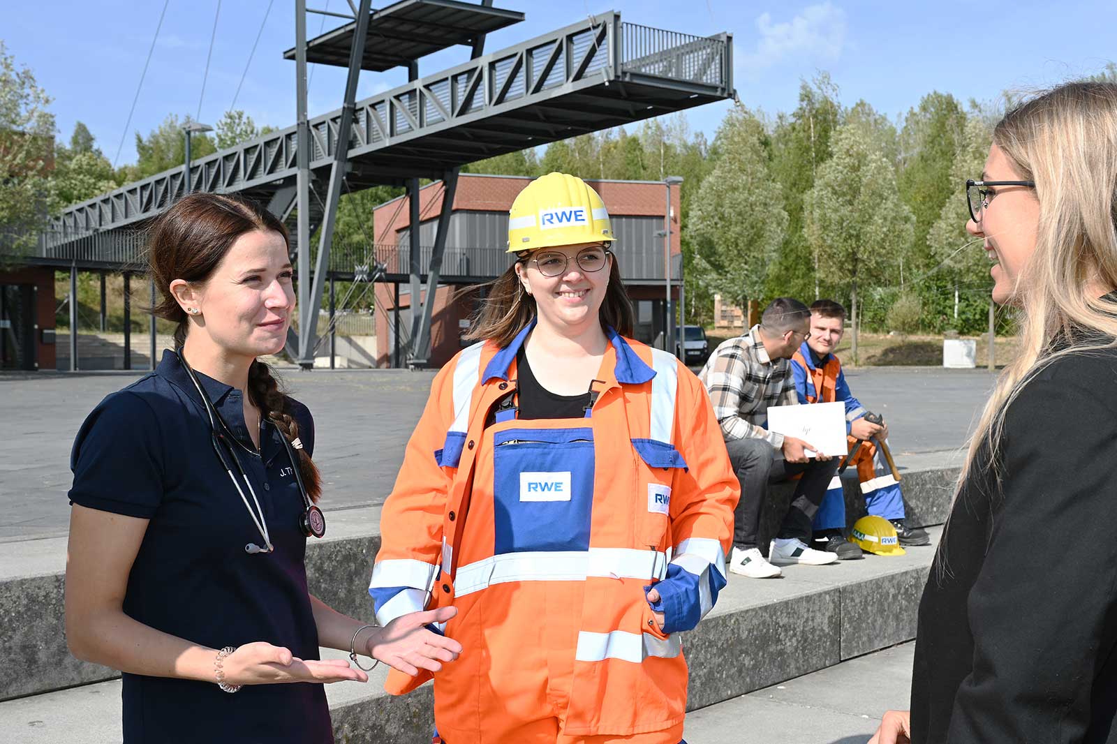 Zwei Frauen führen ein Gespräch, während sie Arbeitskleidung tragen, im Hintergrund sitzen Menschen an einem Außenstandort.