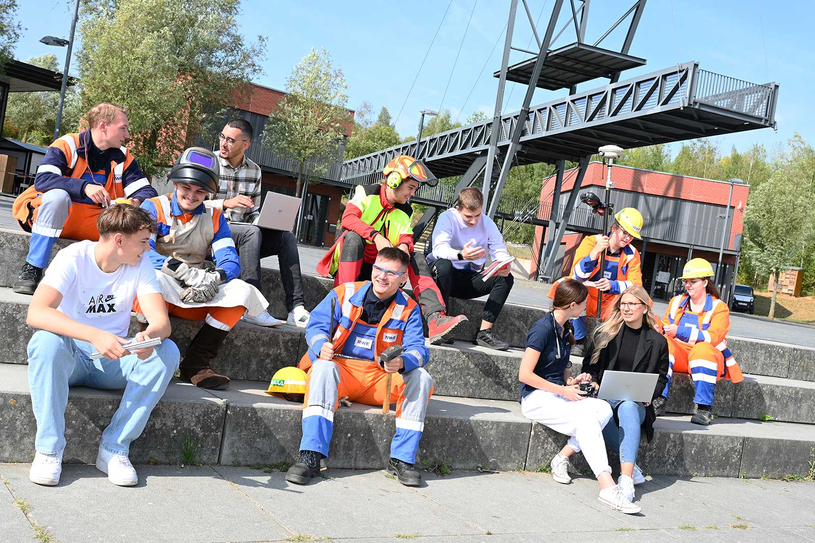 Eine vielfältige Gruppe von Personen in Arbeitskleidung sitzt auf Stufen und nutzt Laptops und Tablets in einer Außenumgebung.