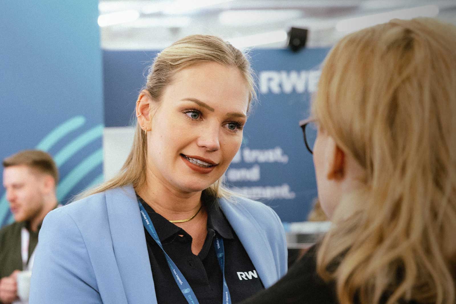 A woman in a blue blazer speaks with another person at a professional event, with branded materials visible in the background.