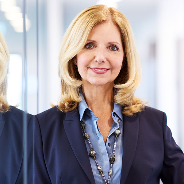 Eine Frau mit blonden, welligen Haaren in einem eleganten Anzug steht vor einem Spiegel und trägt eine Perlenkette. Porträt von Katja van Doren.