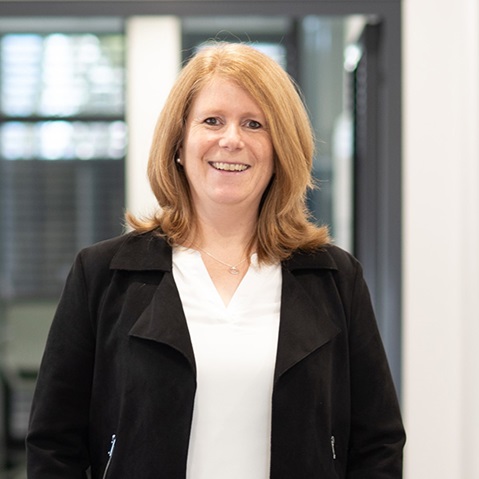 Eine Frau mit blonden Haaren trägt einen schwarzen Blazer und eine weisse Bluse. Der Hintergrund ist eine moderne Büroumgebung.