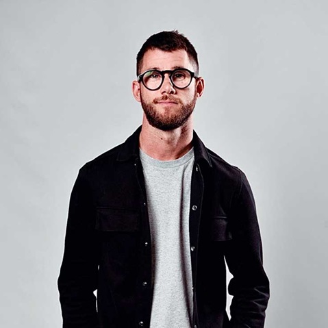 A man wearing a black shirt and a grey T-shirt stands against a neutral background.