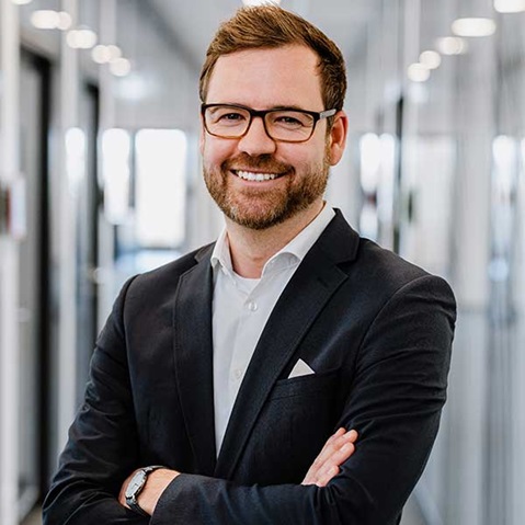 A man in a suit stands with crossed arms in a modern office with glass walls.
