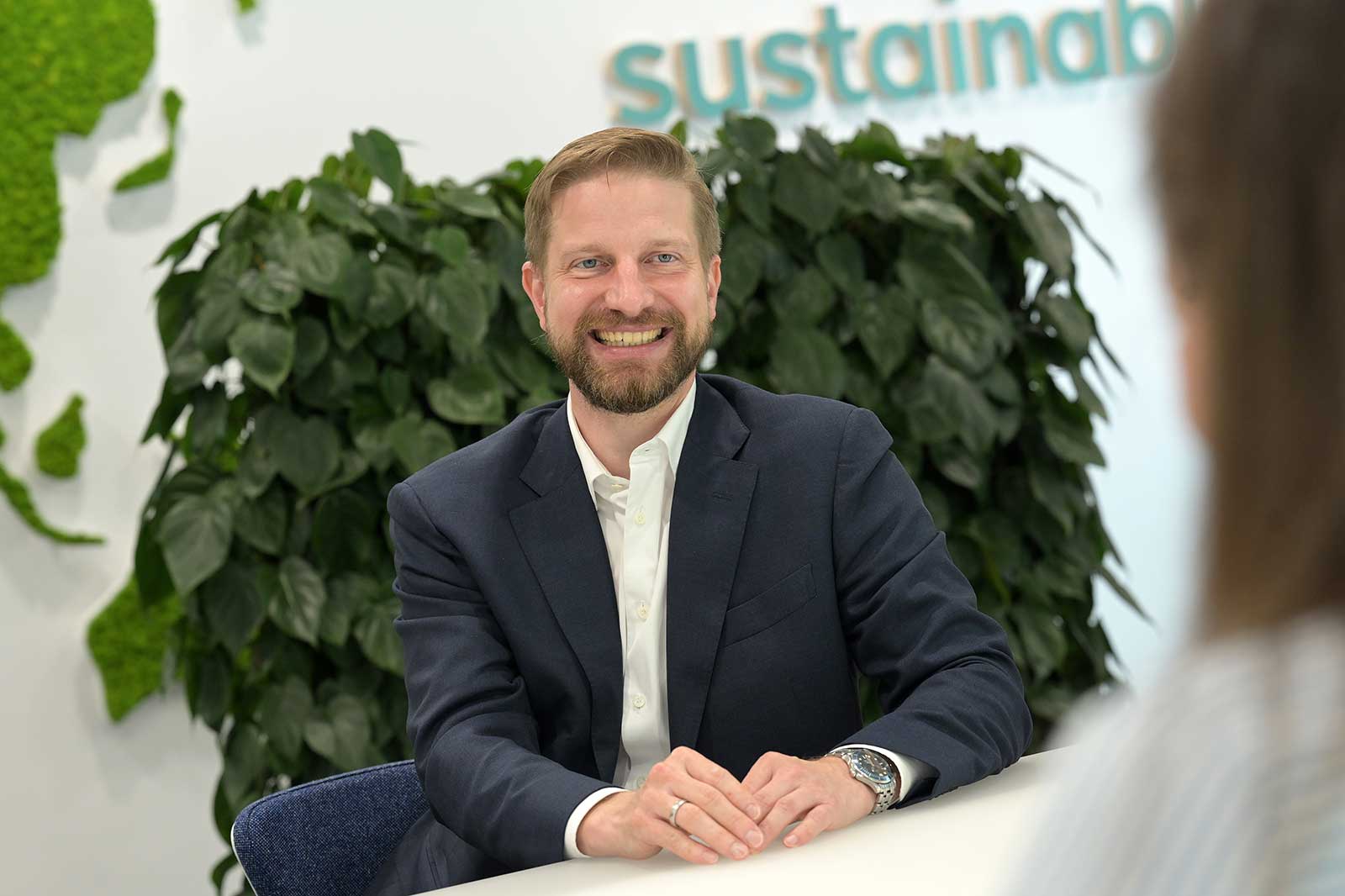 A man in a suit sits at a table with green plants in the background, discussing sustainability.