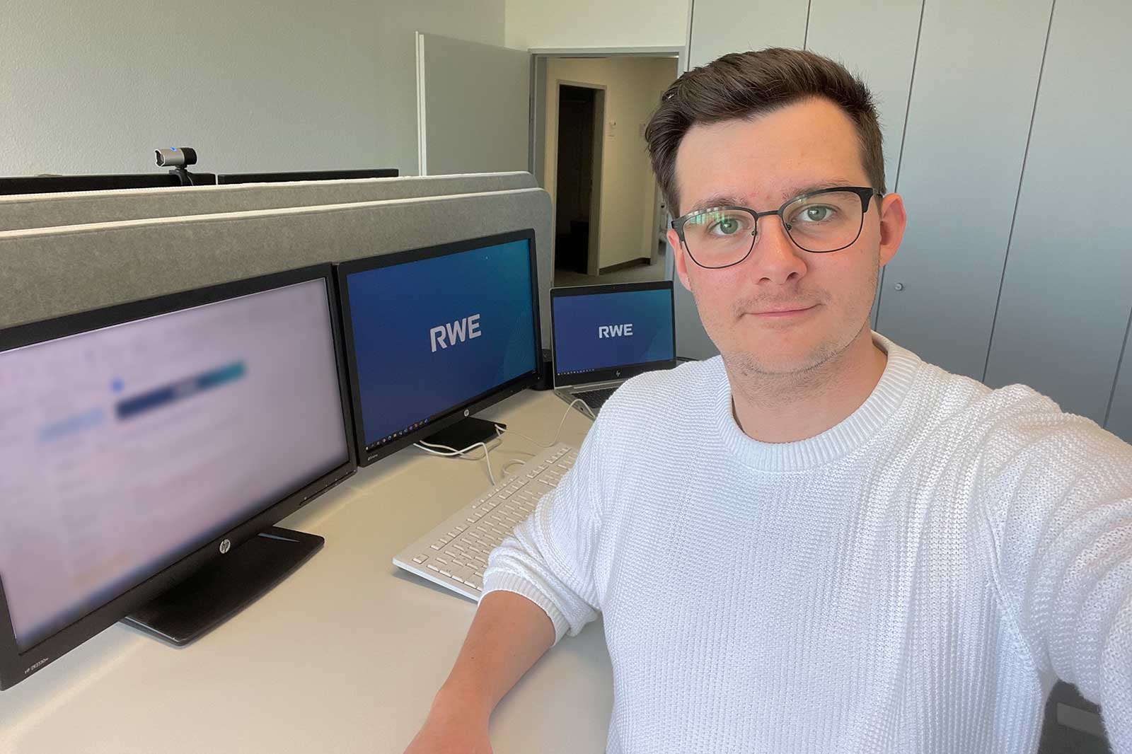 A person in a white sweater poses in an office with three computer monitors displaying the RWE logo.