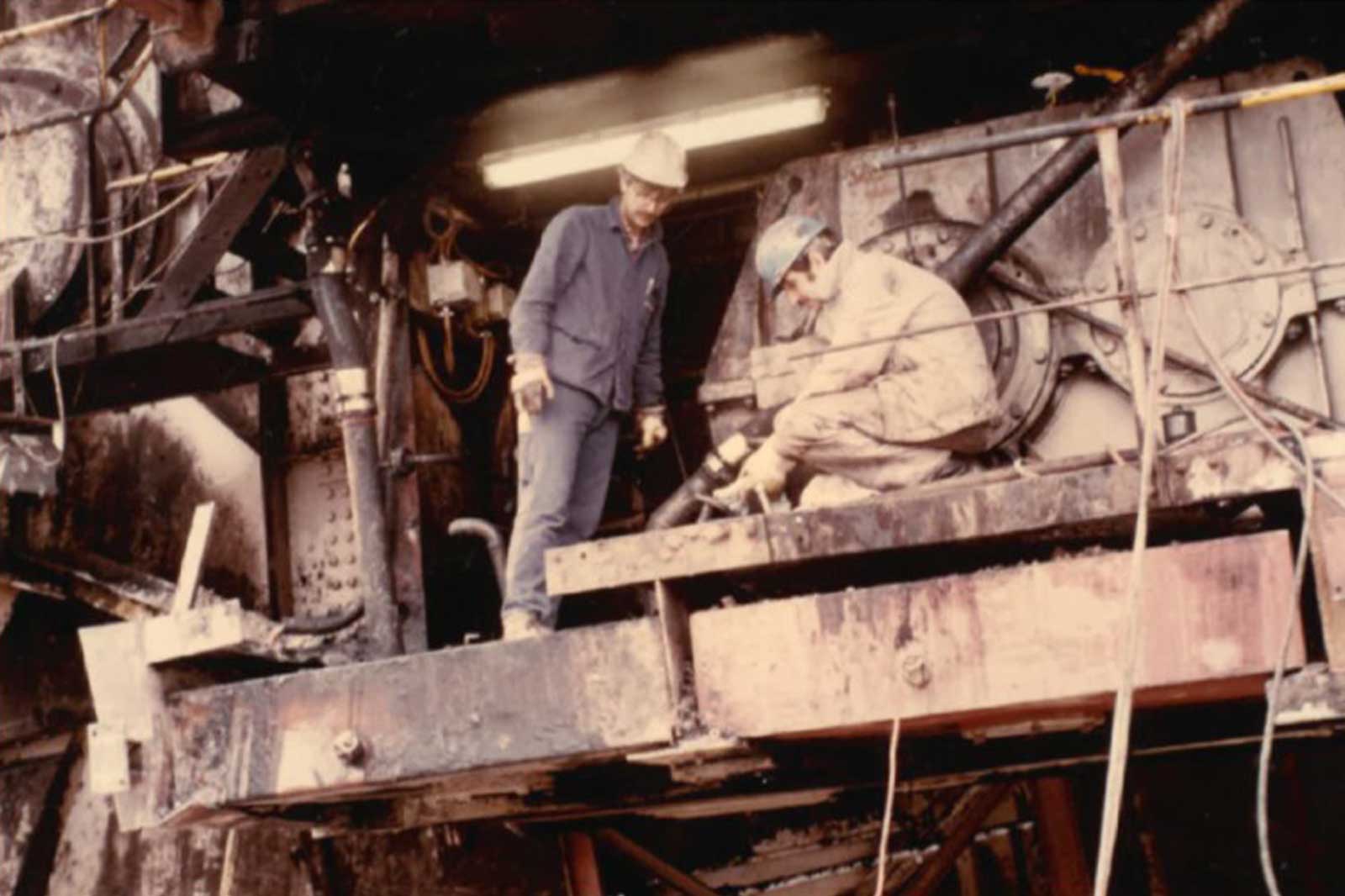 Zwei Arbeiter führen Wartungsarbeiten an schweren Maschinen in einer industriellen Umgebung durch, umgeben von Rohren und Ausrüstung.