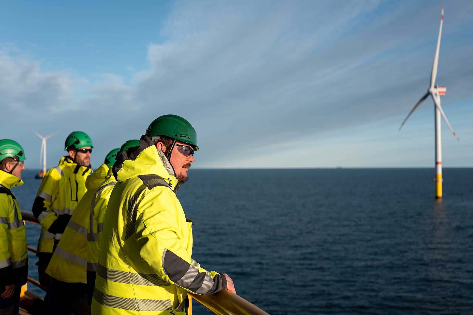 Mehrere Arbeiter in gelben Warnjacken und grünen Helmen stehen auf einer Offshore-Plattform mit unscharfen Gesichtern und blicken auf Windräder im Meer.