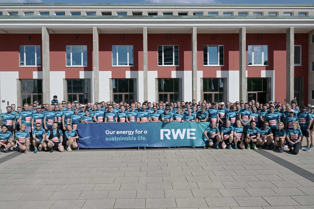 Group in teal shirts pose behind a long blue banner reading "Our energy for a sustainable life." and "RWE" before a red building.