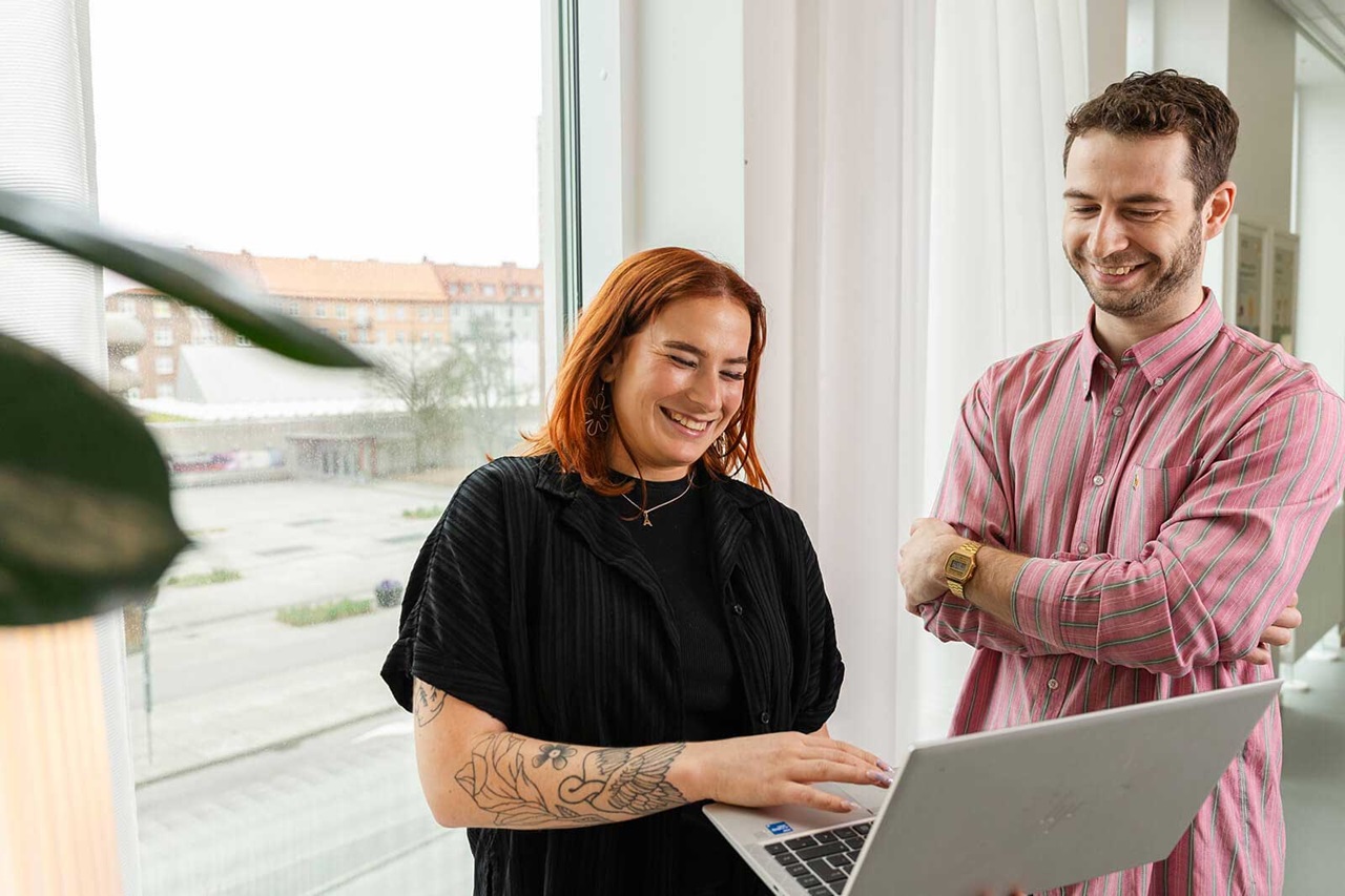 Two people stand by a window, looking at a laptop. The woman has a tattooed arm.