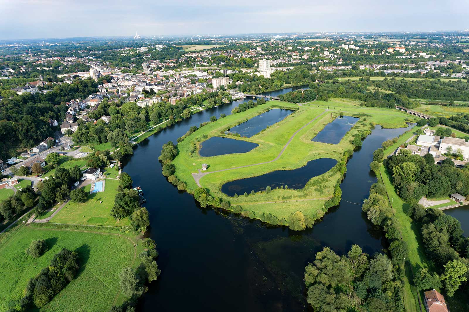 An aerial view of a city featuring green fields, water bodies, and a mix of residential buildings and trees.