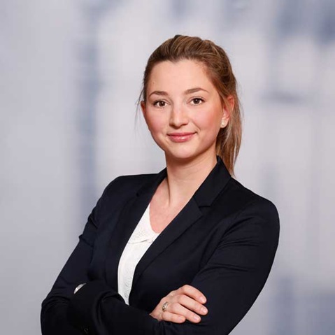 A woman in a black blazer stands with her arms crossed in front of a blurred background.