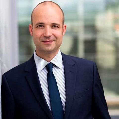 A man in a suit with a tie stands confidently in front of a modern building backdrop.